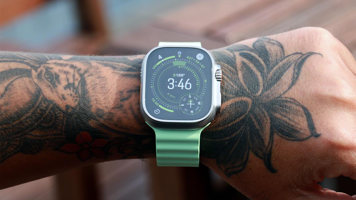 Apple Watch Ultra de titanio con correa Ocean verde claro en una muñeca tatuada, mostrando esfera digital con brújula y hora en tiempo real.