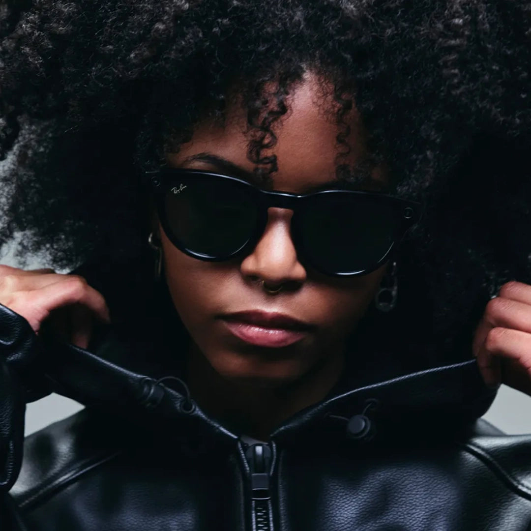 Primer plano de mujer con chaqueta de cuero y pelo rizado usando gafas inteligentes Ray-Ban Meta Wayfarer negras con lentes de sol oscuras.